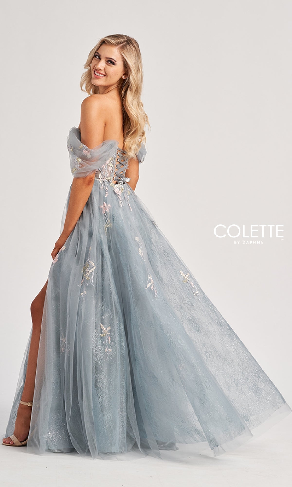 A woman in a light blue, off-the-shoulder tulle gown with floral embroidery and lace-up back smiles sideways, wearing the Long Prom Dress: Colette CL8150. The